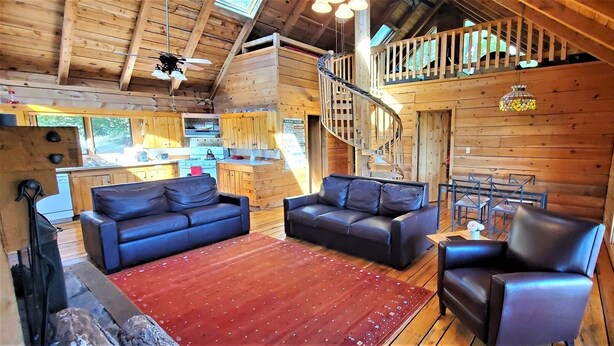 Interior de una cabaña de madera con una sala de estar que incluye dos sofás de cuero negro, un tapete rojo y una escalera que sube a un balcón.