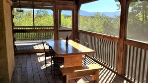Una terraza de madera con una mesa y sillas, con vistas a una zona boscosa y montañas a lo lejos.