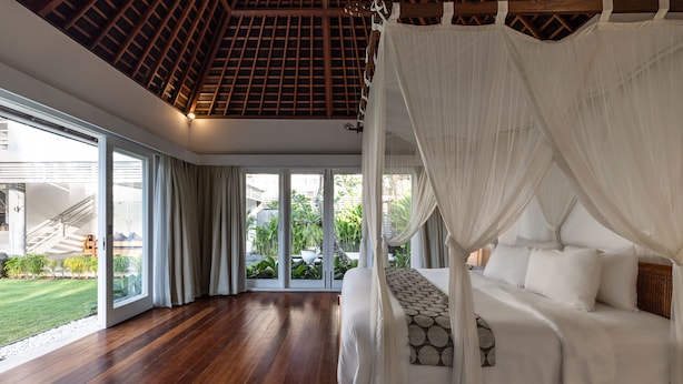 A bedroom with a canopy bed, wooden flooring, and large windows offering a view of the outdoors. - Kerta Rama Seminyak (Keroboken Kelod)