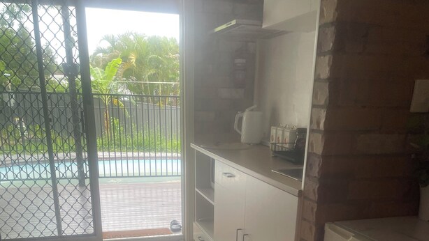 A kitchen with a refrigerator, a coffee maker, and a sink. There is a door leading to a balcony with a view of palm trees and a swimming pool.