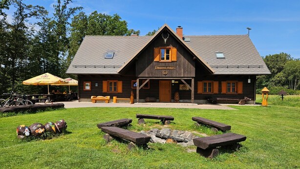 A rustic lodge with a central entrance, wooden benches, picnic tables, and a stone fire pit.