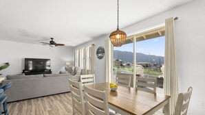 A modern living room with a wooden dining table, white chairs, a sofa, a TV, and a ceiling fan.