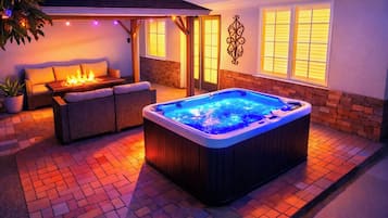 A hot tub with a wooden exterior and a blue interior, surrounded by a brick patio and a covered seating area with cushions.