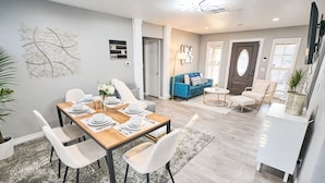 A modern dining area with a wooden table set for four, surrounded by white chairs. There is a blue sofa and a small round table in the background. - Modern Retreat w/ Private Spa, Arcade, Playground, Disney, Convention & Beaches (Anaheim)