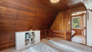Un dormitorio acogedor en el ático con paredes de madera, un aparador blanco lleno de libros, una cama y una ventana con cortinas.