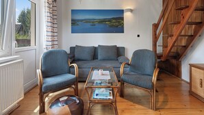Una sala de estar acogedora con un sofá, dos sillones, una mesita de centro y una lámpara colgante. Un ventanal ofrece vistas a la vegetación exterior.