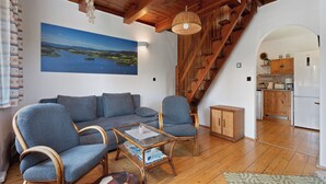 Un salón acogedor con techo de madera, un sofá, dos sillones, una mesa de centro y vistas a una cocina y una escalera.