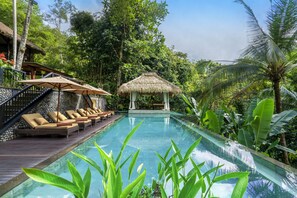 Un espace piscine doté de chaises longues et d’un gazebo à toit de chaume, le tout entouré d’une végétation luxuriante.