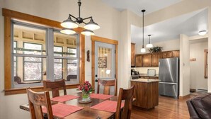 Un comedor con mesa de madera, sillas y una araña. Se observa una cocina al fondo con armarios de madera y un refrigerador de acero inoxidable.