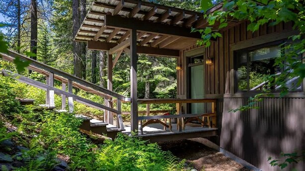 Una cabaña de madera con un porche cubierto, una escalera y vistas del bosque circundante.