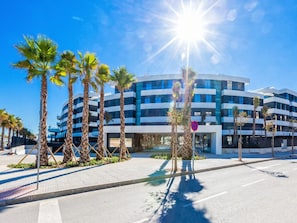 A modern multi-story building with a curved facade, surrounded by palm trees and a clear blue sky. - Galatea | Beachfront - pool - gym (Torremolinos)