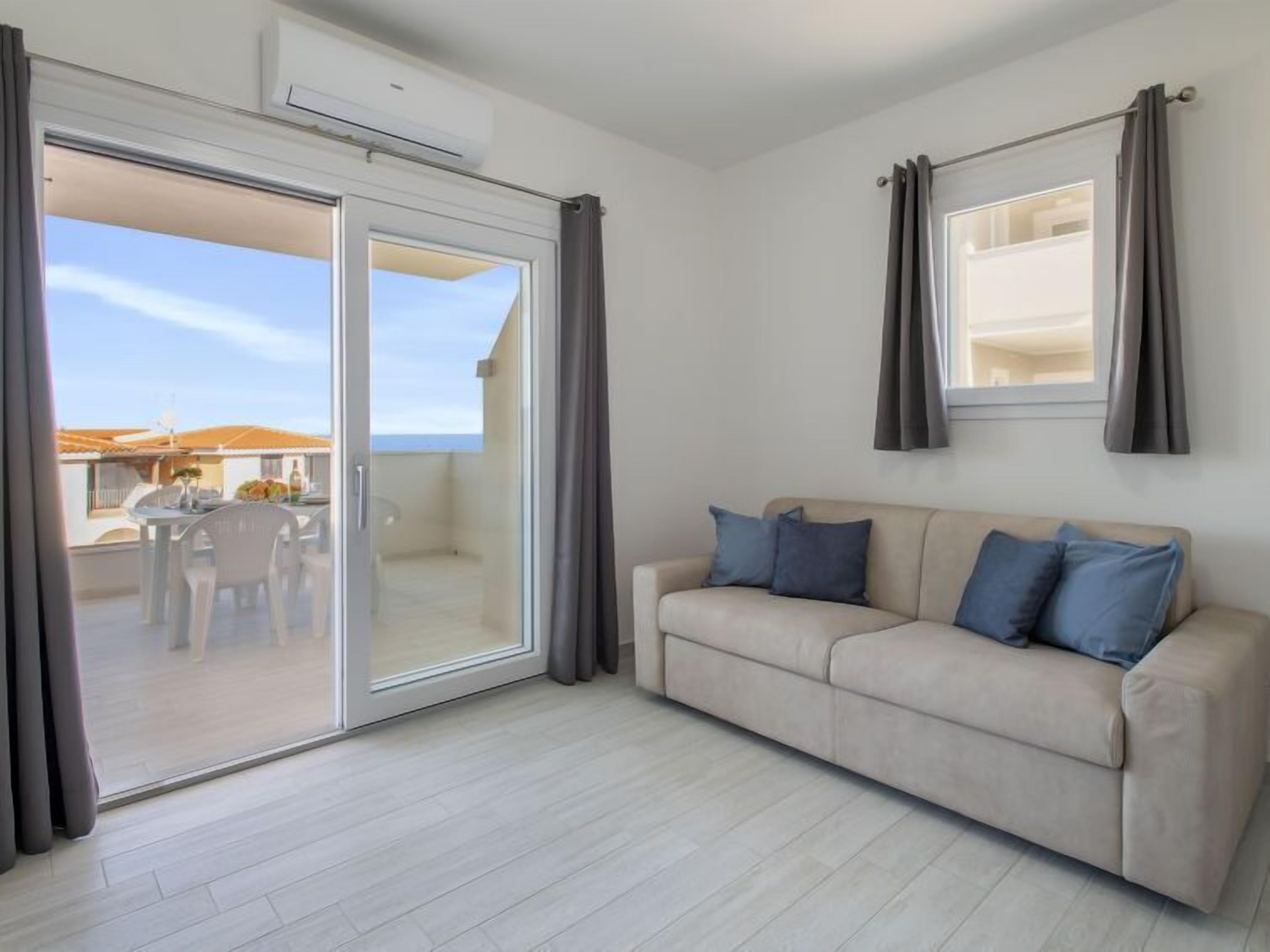 A modern living room with a sliding glass door leading to a balcony, a beige sofa with blue cushions, and a small table with chairs.