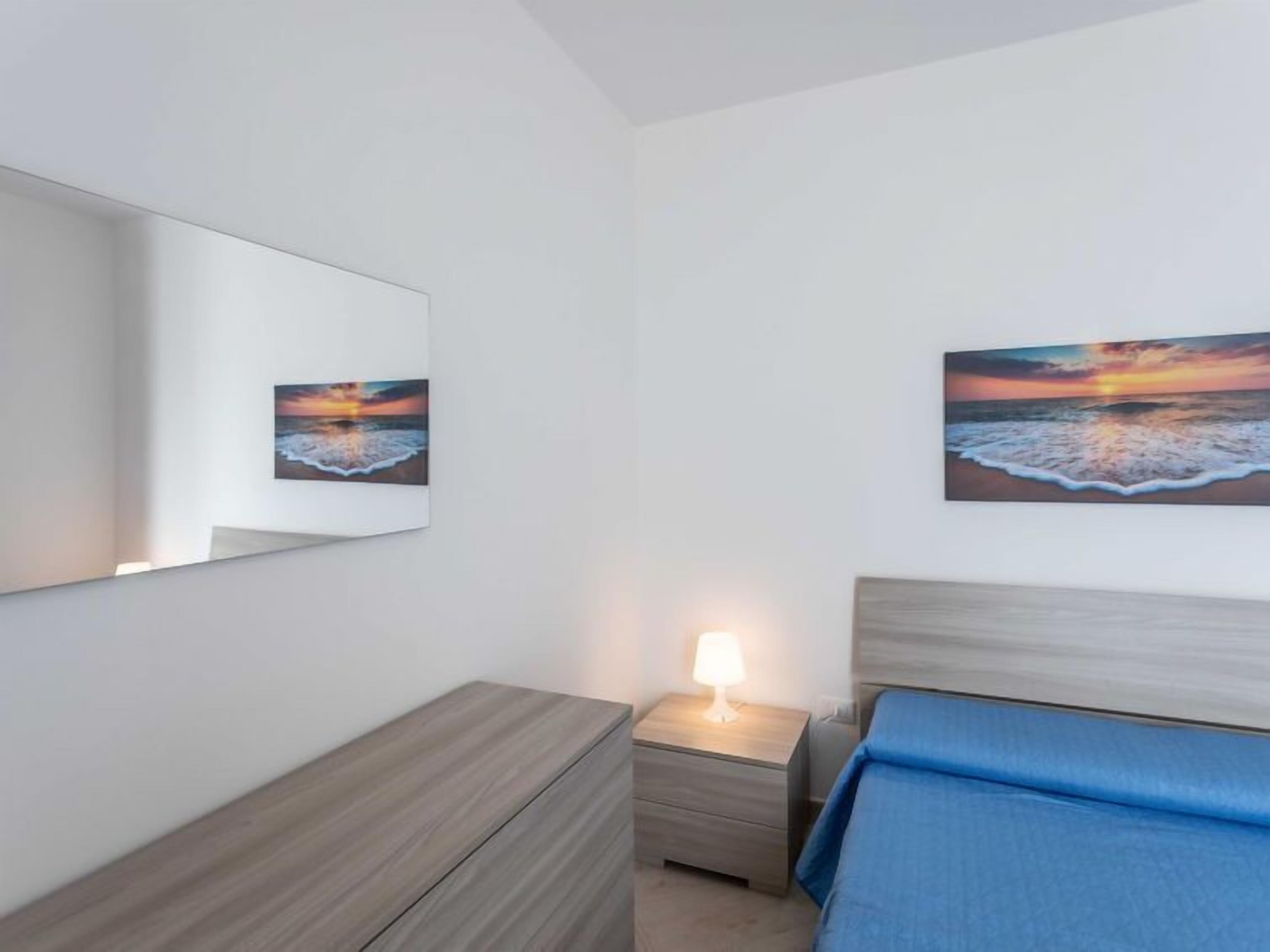 Ein Schlafzimmer mit einem Bett, einem Nachttisch mit Lampe, einer Kommode und zwei Bildern eines Sonnenuntergangs über dem Meer.