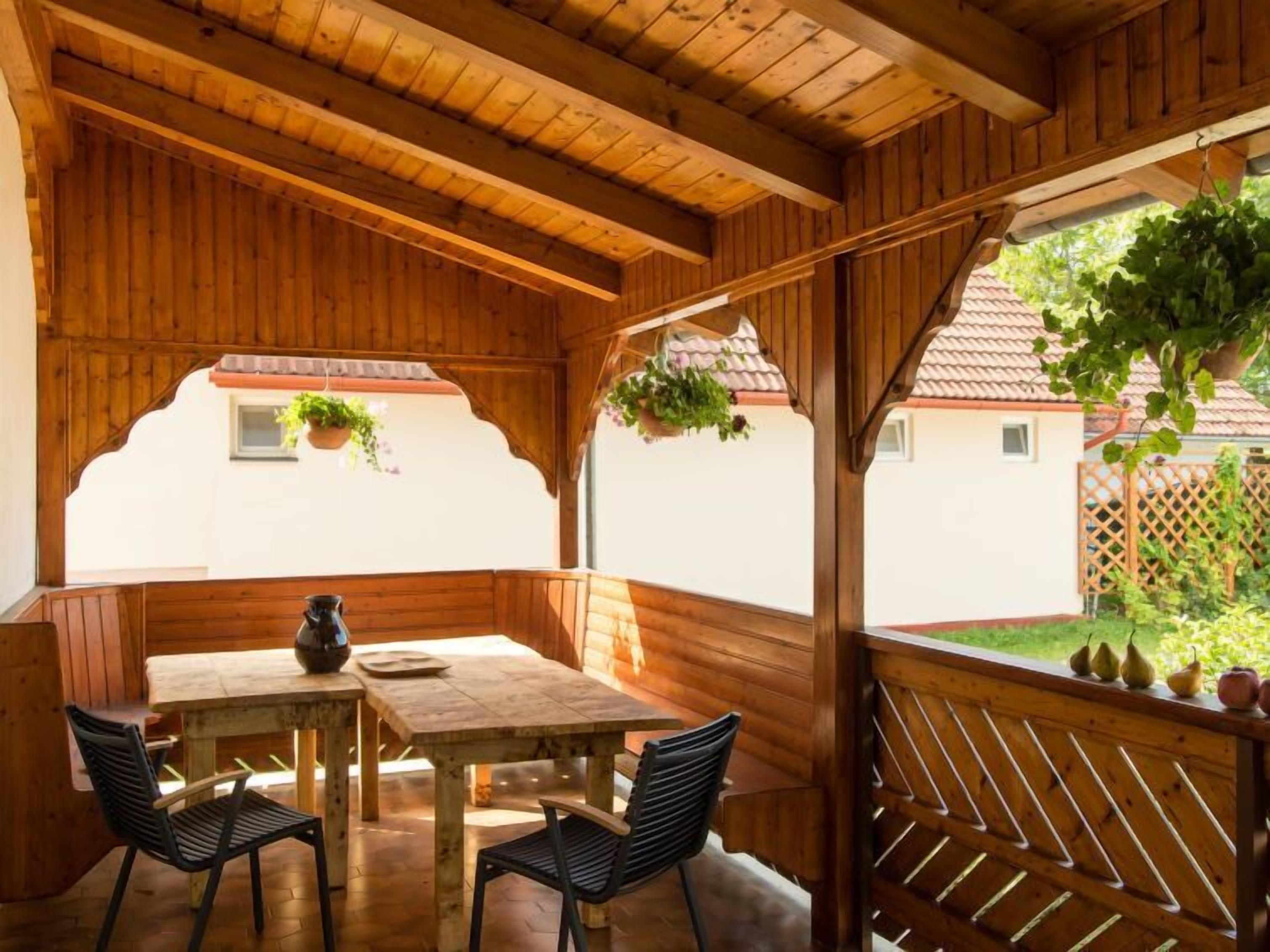 Un área de comedor al aire libre con madera, que incluye una mesa, sillas, una planta colgante y vistas a una casa y jardín.