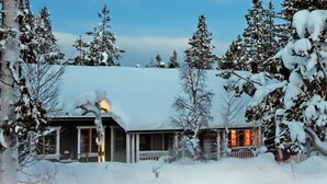 Une cabane enneigée, illuminée de l’intérieur par une lumière chaleureuse, entourée d’arbres chargés de neige.