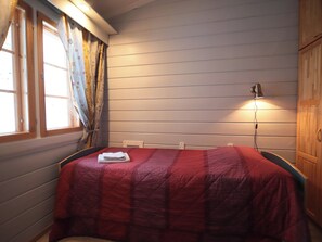Une chambre à coucher avec un lit, une fenêtre avec des rideaux, une lampe fixée au mur et une armoire en bois.