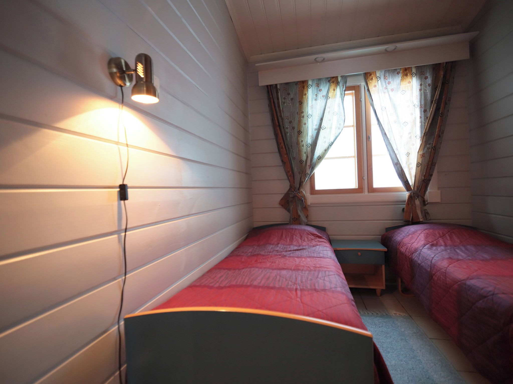 A room with two single beds, a small table, a wall-mounted light, and a window with curtains.