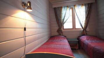 A room with two single beds, a small table, a wall-mounted light, and a window with curtains.