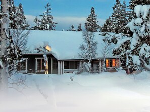 Un chalet enneigé, doté d’un porche bien visible et d’une fenêtre éclairée, entouré d’arbres.