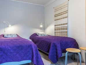 Une chambre avec deux lits, chacun recouvert d’une couverture violette et agrémenté d’un oreiller blanc. On y trouve également un petit tabouret et une lampe fixée au mur.
