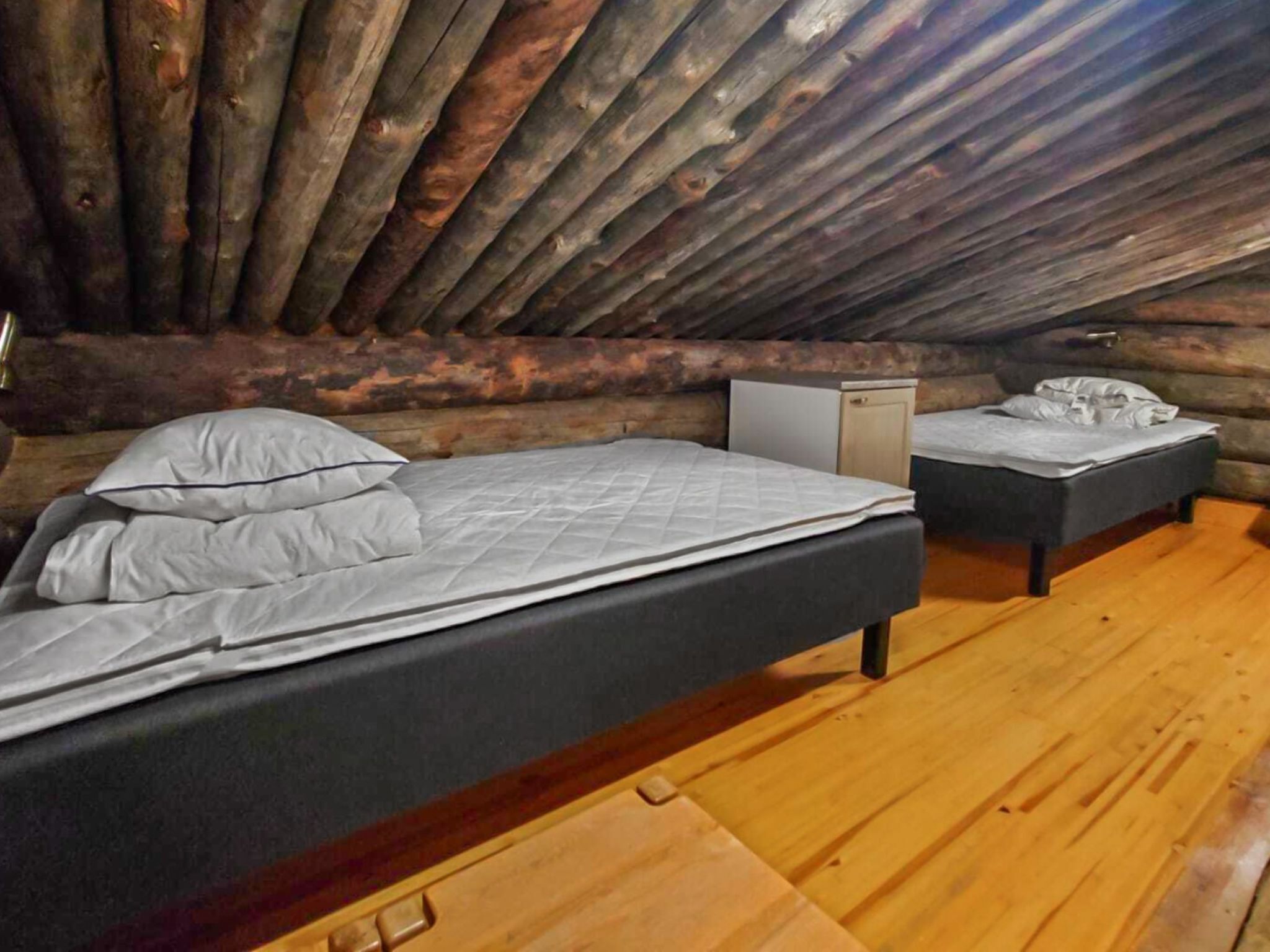 Un dormitorio tipo cabaña de madera con dos camas, un pequeño aparador blanco y suelo de madera.