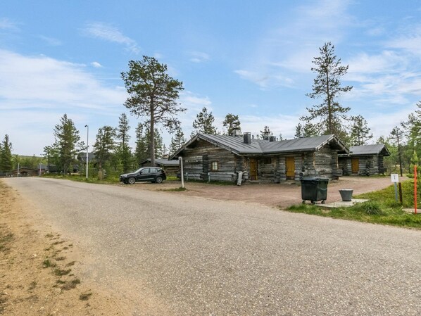 Une cabane rustique en rondins de bois, avec une allée en gravier, un VUS garé et un ciel dégagé.
