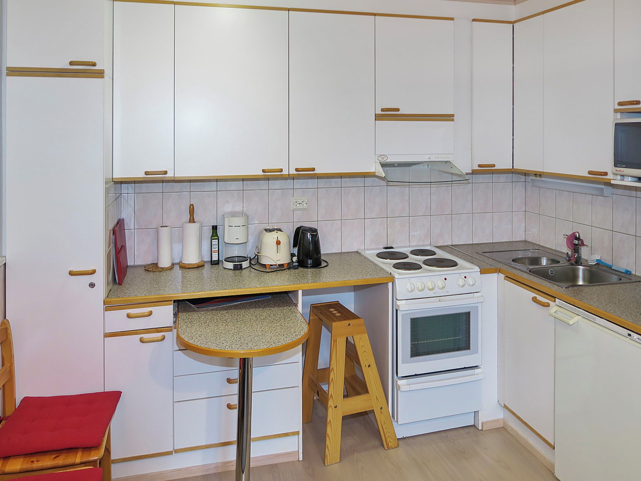 A compact kitchen with white cabinets, a stove, a sink, and a wooden stool.