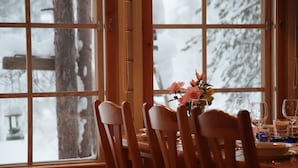 Ein Essbereich mit Holzkästen und einem Tisch, einer Vase mit Blumen und Blick auf schneebedeckte Bäume durch große Fenster.