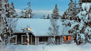 Ein mit Schnee bedecktes Häuschen mit warmem Licht im Inneren, umgeben von schneebedeckten Bäumen.