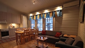 Ein gemütliches Wohnzimmer mit einer Couch, einem Couchtisch und zwei Hocker. An das offene Wohnzimmer grenzt ein Essbereich mit Tisch und Stühlen, der an eine Küche angrenzt, die einen Herd und Schränke beinhaltet. Große Fenster mit Vorhängen sorgen für Tageslicht und Blick ins Freie.