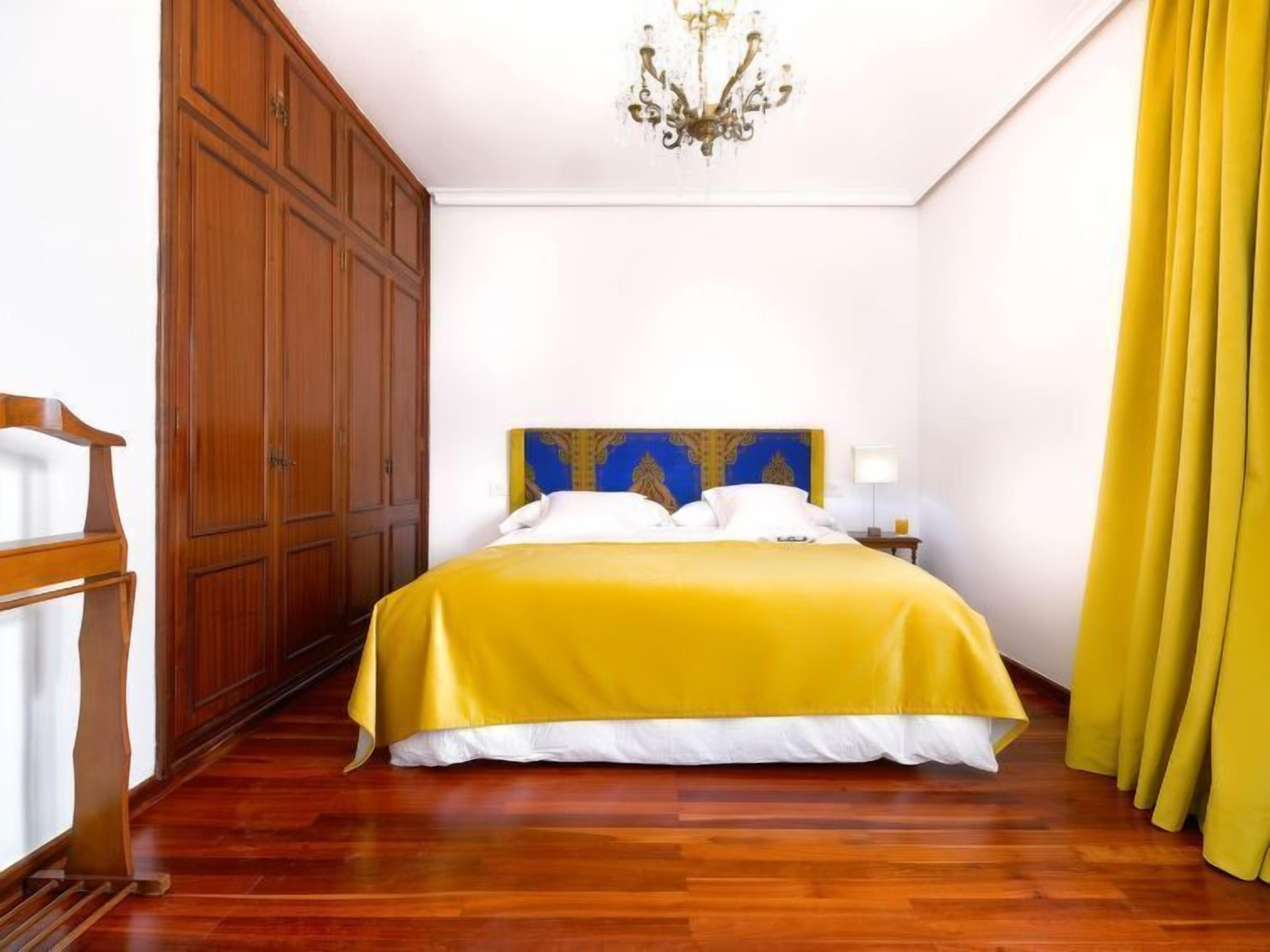 A hotel room with a wooden wardrobe, a bed with a yellow bedspread, a chandelier, and yellow curtains.