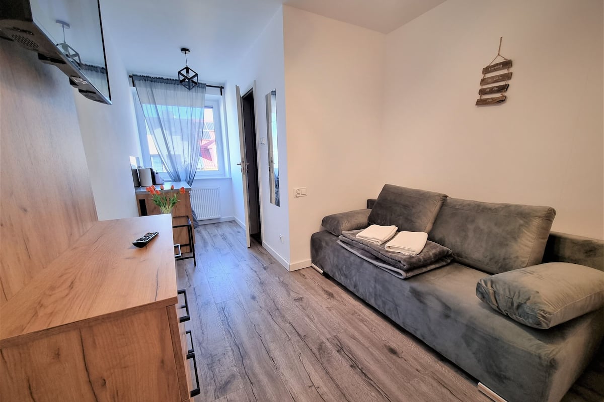 Un salon moderne avec un canapé gris, une table basse en bois et un petit coin cuisine doté d’une fenêtre et de rideaux.