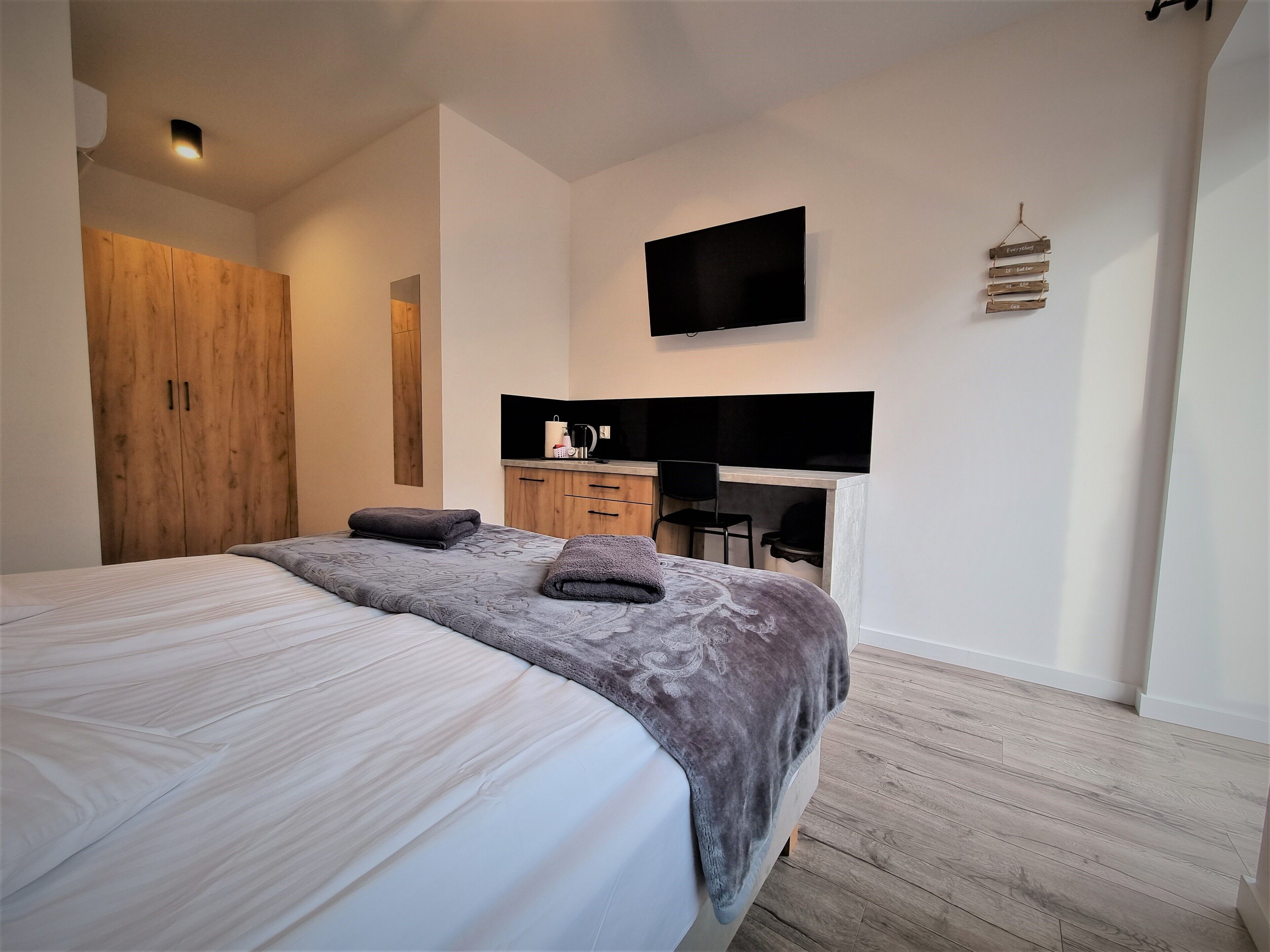 A bedroom with a bed, a wooden wardrobe, a desk with a chair, a flat-screen TV, and a wall-mounted shelf with decorative items.