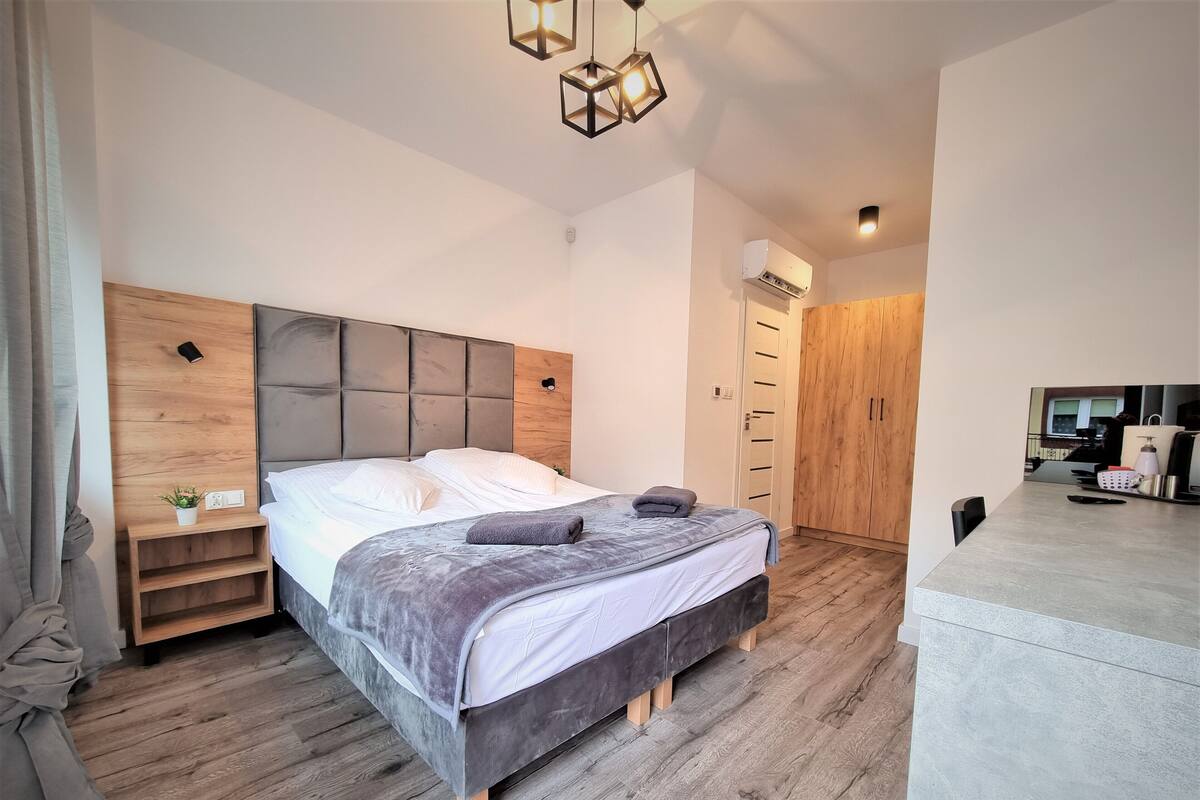Une chambre moderne avec une tête de lit en bois, un lit avec une literie blanche et grise, une table de chevet avec une lampe et une armoire.