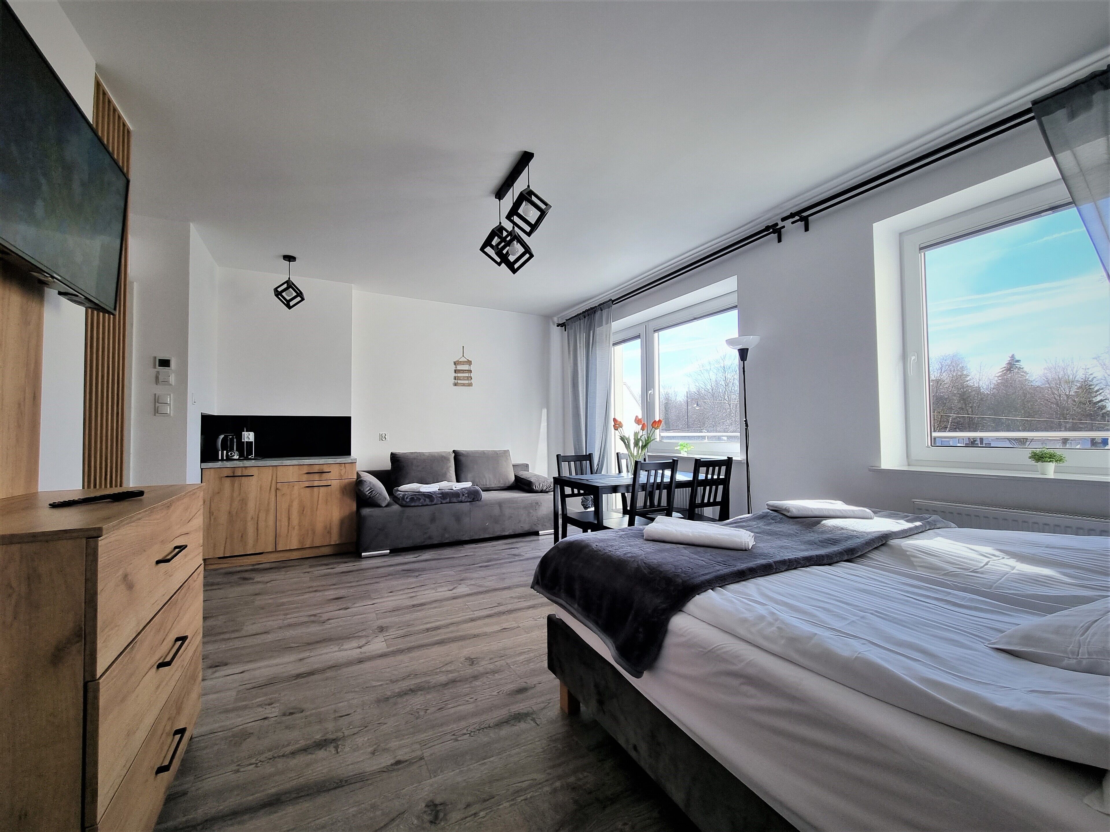 Un dormitorio moderno con cama, cómoda de madera, televisor, mesa de comedor con sillas y ventana con vista a árboles.