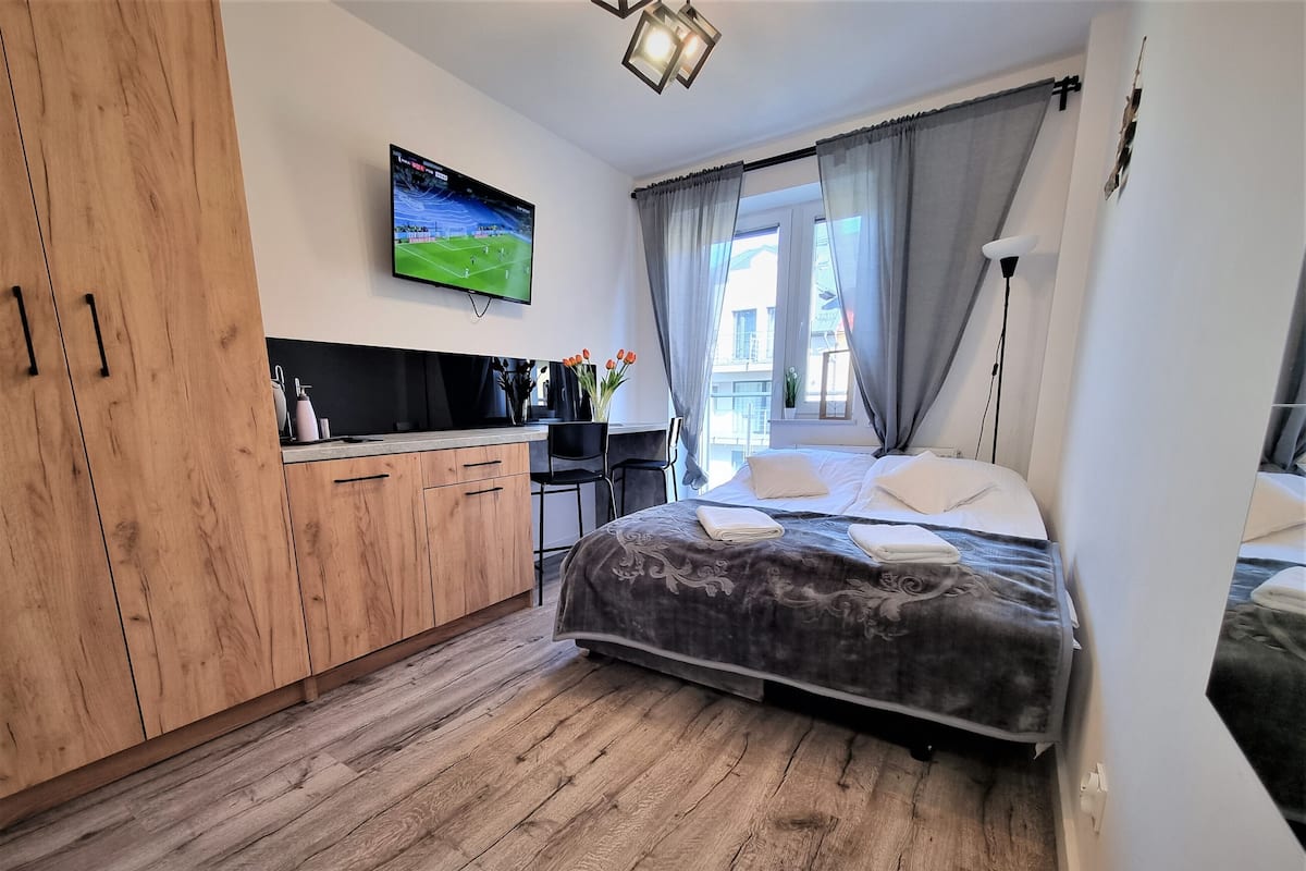 Une chambre moderne avec un lit, un téléviseur fixé au mur, un plancher en bois et une fenêtre avec des rideaux.