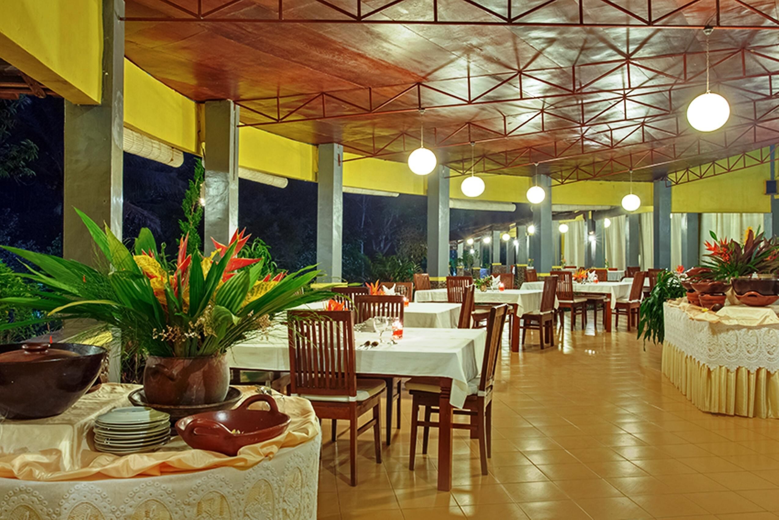A restaurant with multiple tables and chairs, large potted plants, and a tiled floor.