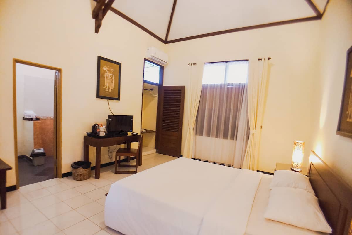 A hotel room with a bed, a desk with a computer, an air conditioning unit, a window with curtains, and a bedside lamp.