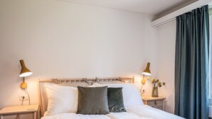 Una cama bien hecha con sábanas blancas, dos lámparas de noche, una mesita de noche con un florero y una ventana con cortinas.