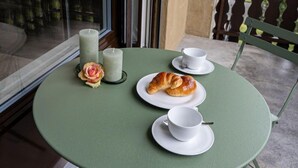 Una mesa redonda con mantel verde, dos platos blancos con cruasanes, dos tazas blancas y dos velas en un balcón con vistas a la vegetación.