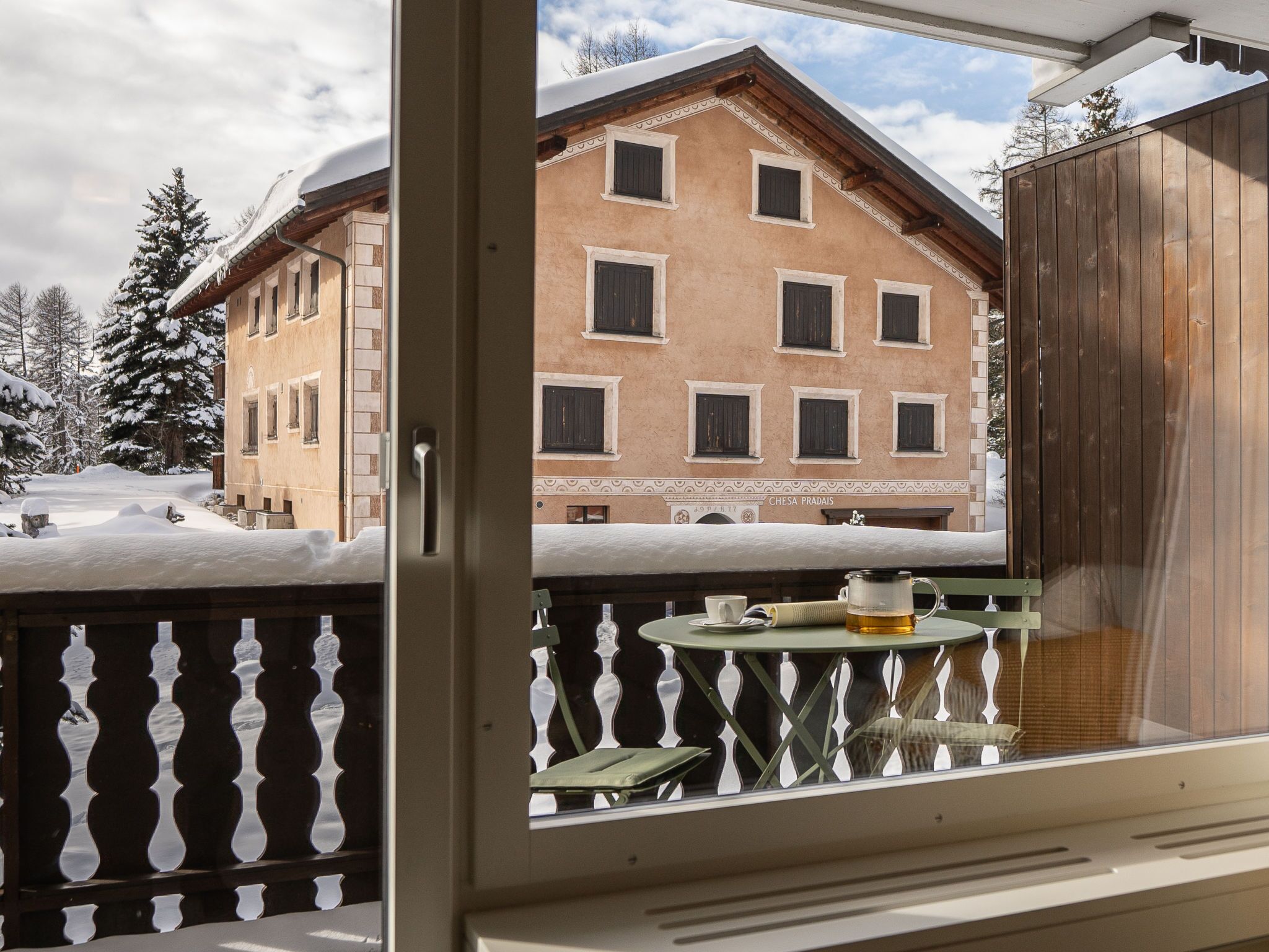 A balcony with a view of a snow-covered building with multiple windows and a wooden fence.