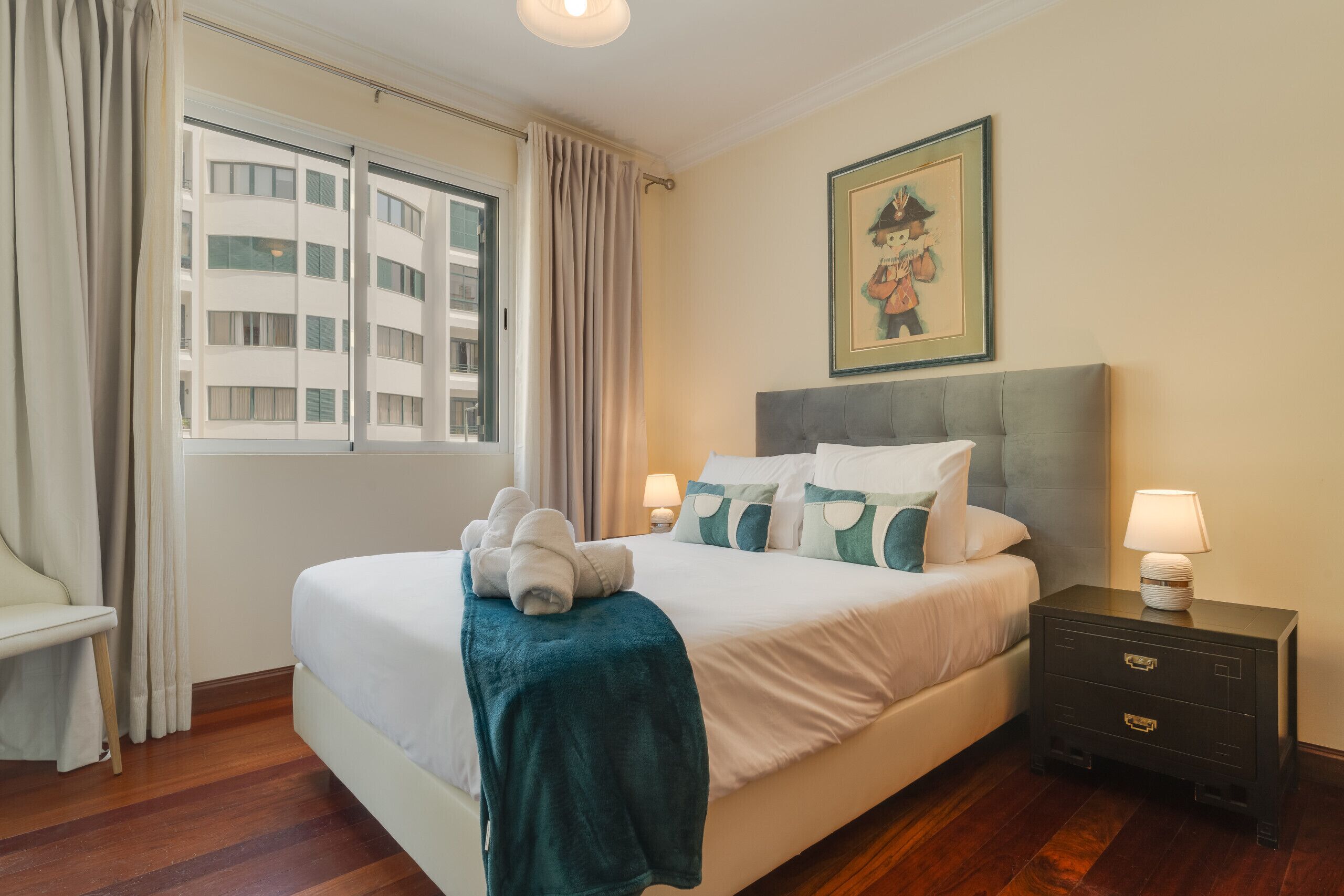 Cama bem arrumada com manta e almofadas verde-água, mesa de cabeceira com abajur e janela com cortinas.