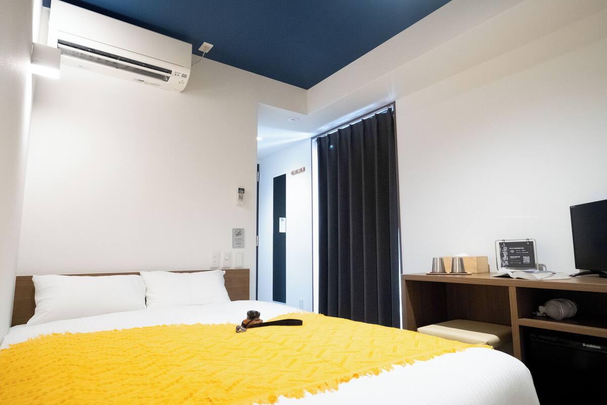 A hotel room with a bed, an air conditioning unit, a desk with a computer, and a television.