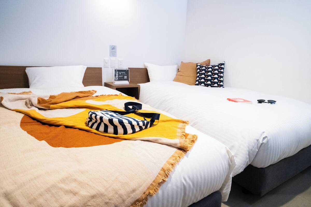 A hotel room with two beds, a nightstand with a radio, and a wall-mounted thermostat.