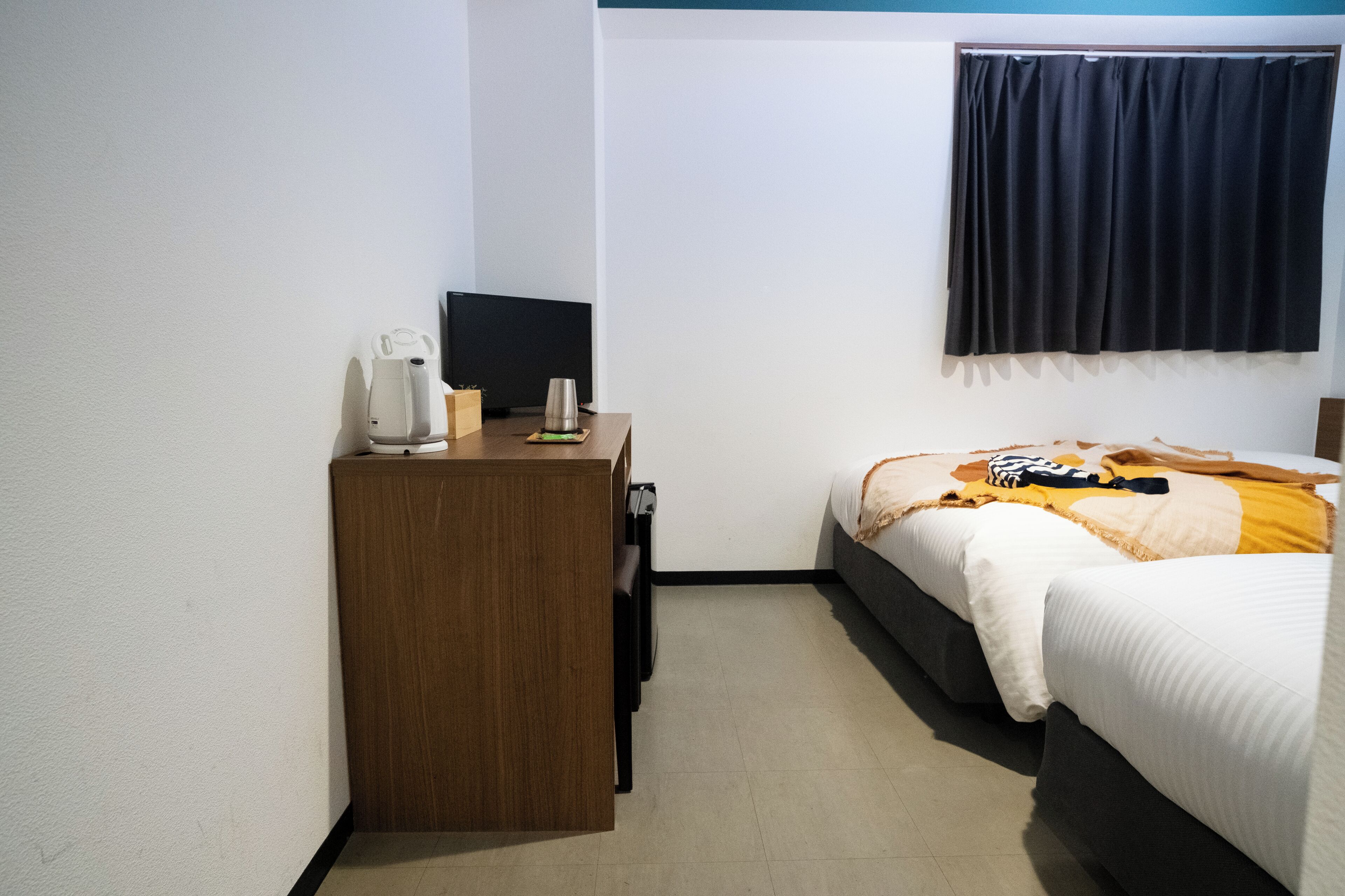 Una habitación de hotel pequeña con un escritorio de madera, una cama con manta blanca y amarilla, y una cortina oscura sobre la ventana.