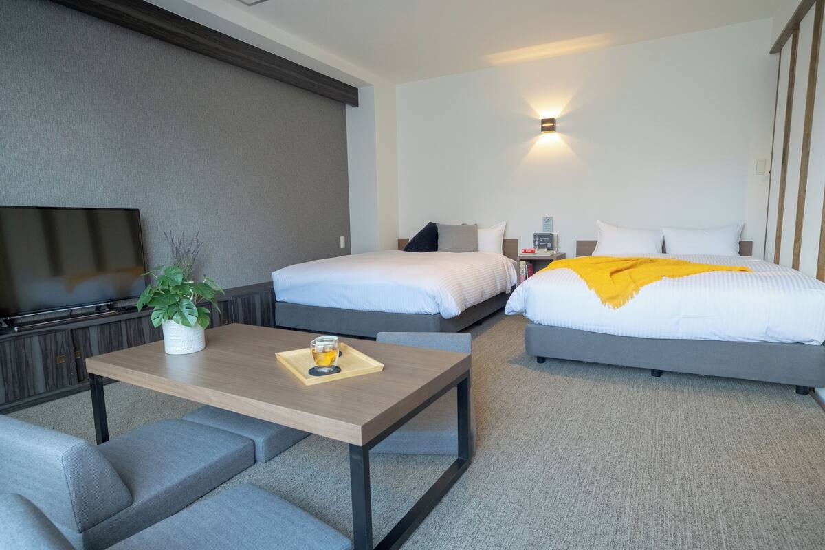 A hotel room with two beds, a TV, a small table with a plant, and a bedside table with a lamp.