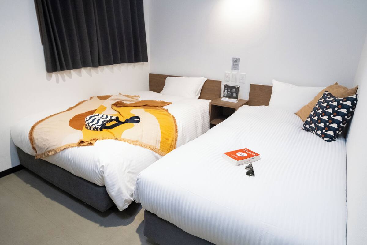 A hotel room with two beds, each with white bedding and different patterned throws. There is a bedside table with a lamp and a small clock.