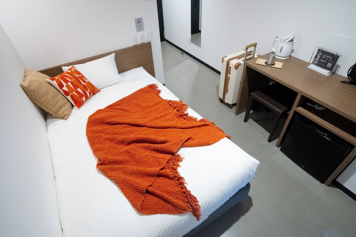 A hotel room with a bed, a desk with a computer, a kettle, and a small refrigerator.