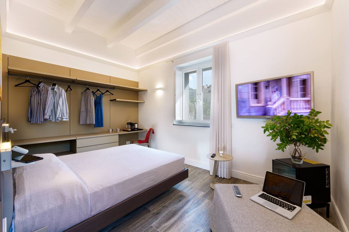Una habitación de hotel moderna con cama, televisión, ordenador portátil, planta y ventana con cortinas.