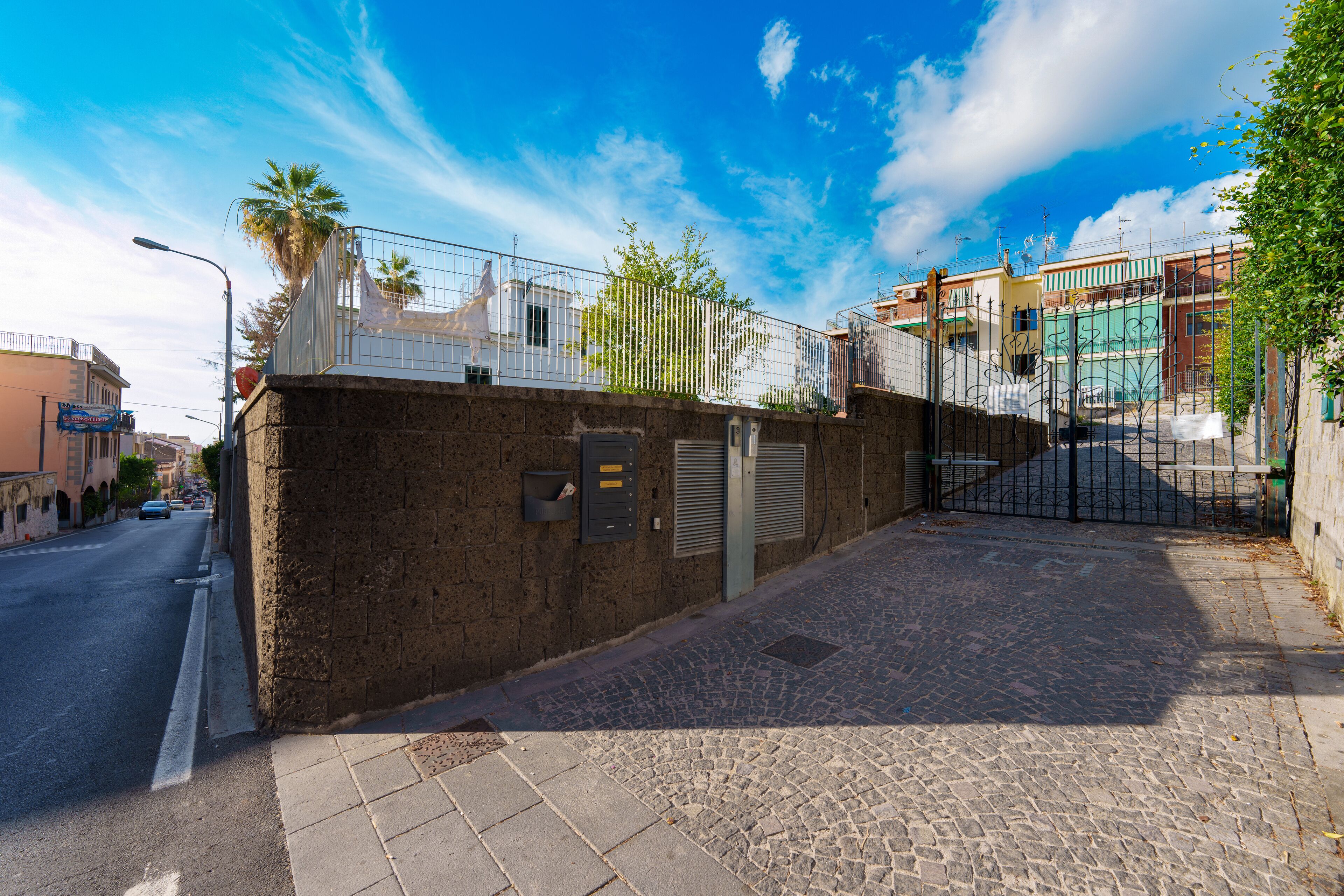 A cobblestone street with a brick wall and metal gate, leading to a building with a balcony and a palm tree.